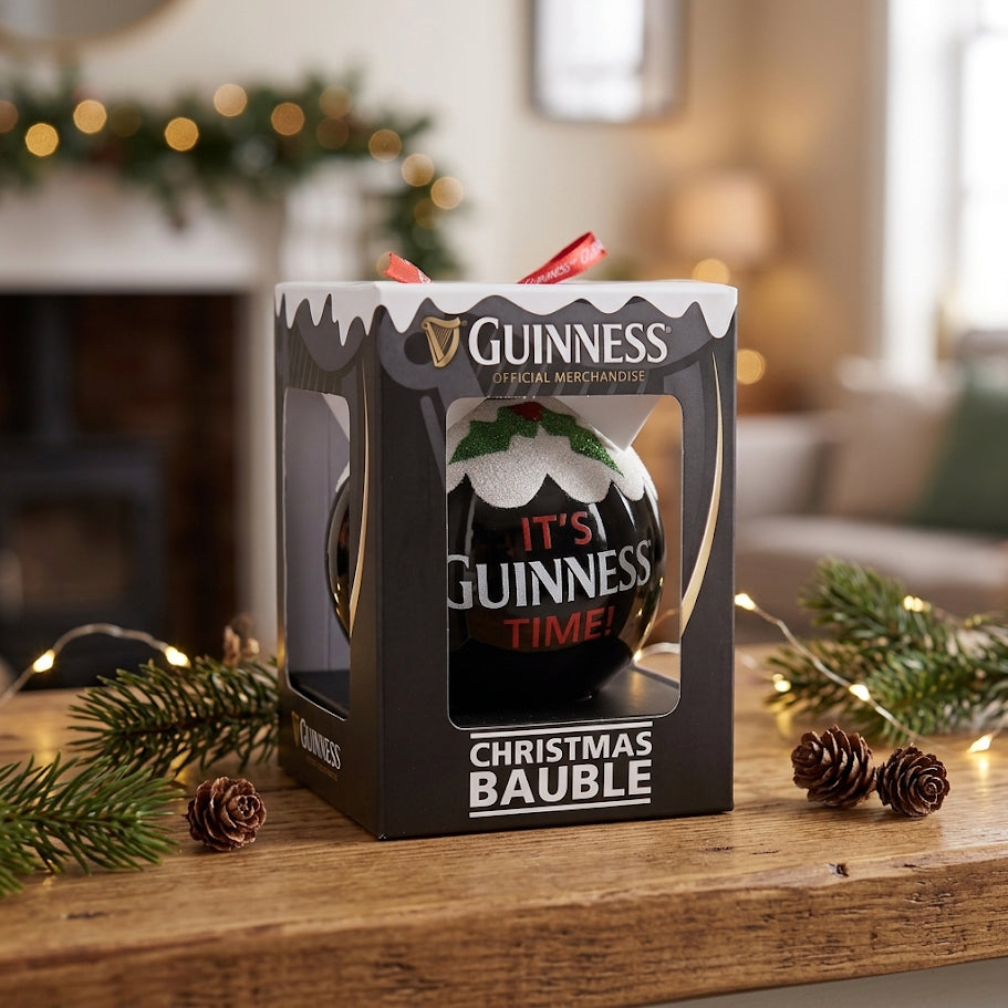 A boxed Guinness Christmas Holiday Pudding Bauble sits on a wooden table with pine branches and fairy lights in the background, making for a charming tree decoration.