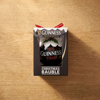 A boxed Guinness Christmas Holiday Pudding Bauble rests on a wooden surface. This festive ornament from Guinness is decorated with holly and a snow-like design on top, along with the slogan “IT’S GUINNESS TIME!”, making it the perfect tree decoration for any fan of the iconic stout.