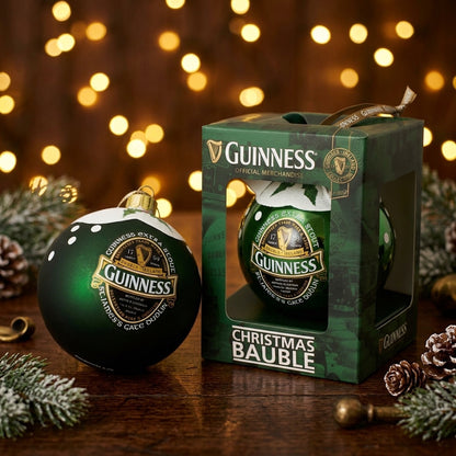 A Guinness Green Collection Gloss Bauble sits next to its box on a wooden table, surrounded by pine cones and holiday lights in the background.