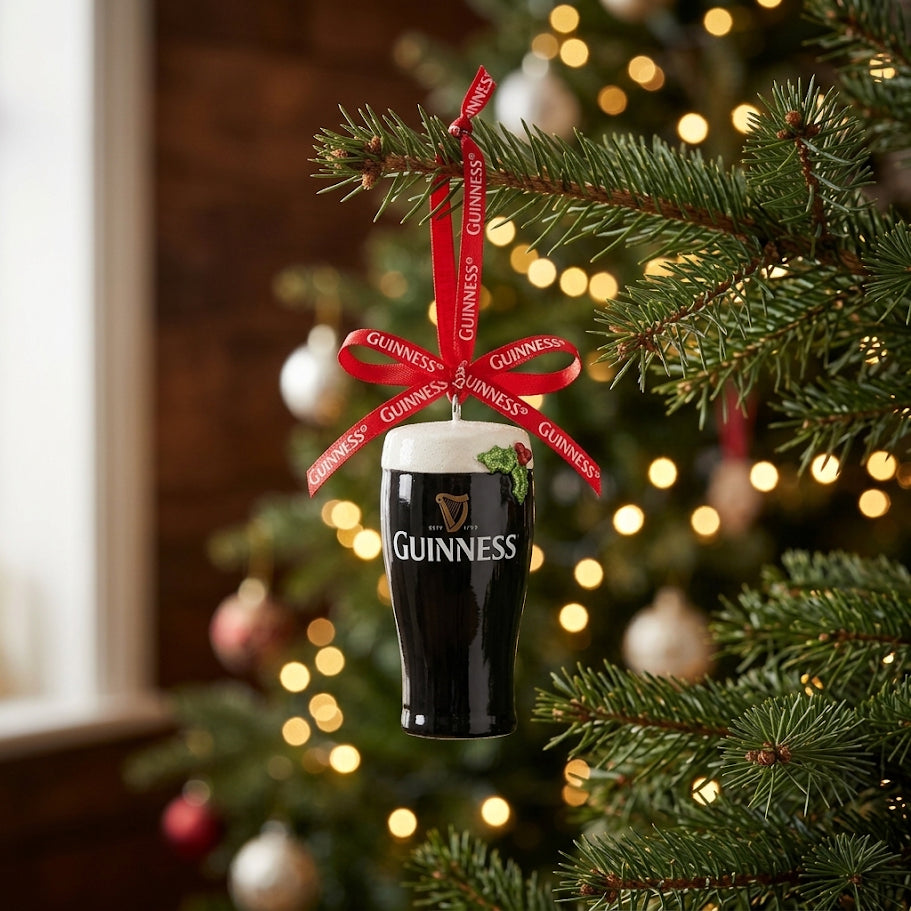 The Christmas Pint Decoration by Guinness hangs on a Christmas tree, adorned with a red Guinness ribbon and surrounded by softly blurred lights, making it the perfect addition to your collection of Guinness Christmas decorations.