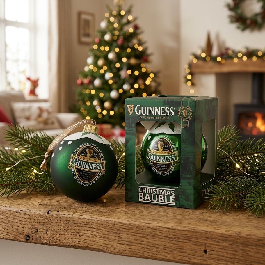 A Green Collection Gloss Bauble by Guinness rests on a wooden mantel beside its box, surrounded by festive decorations and a glowing Christmas tree in the background—a charming nod to the holiday spirit from Guinness.