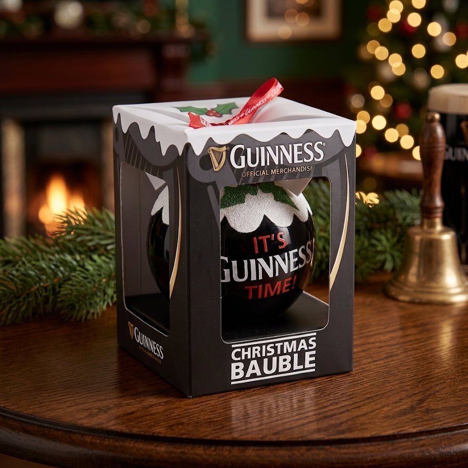 A boxed Guinness Christmas Holiday Pudding Bauble sits on a wooden table, surrounded by festive decorations, with a fireplace and Christmas tree in the background—the perfect tree decoration for fans of Guinness.