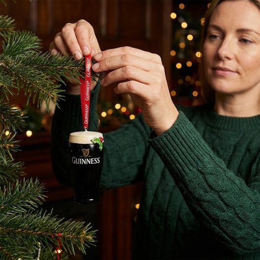 A woman hangs the Guinness Christmas Pint Decoration on a Christmas tree branch, with festive resin Christmas ornaments and holiday accents visible in the background.