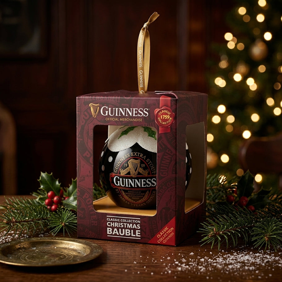 A Guinness Classic Label Glass Bauble from Guinness, presented in a decorative box, rests on a festive table adorned with holly, with a Christmas tree softly blurred in the background—making it the perfect ornament gift for any beer lover this holiday season.