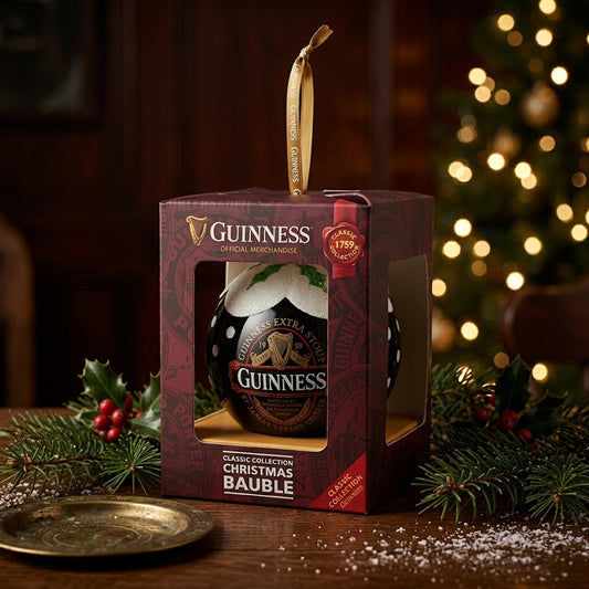 A Guinness Classic Label Glass Bauble from Guinness, presented in a decorative box, rests on a festive table adorned with holly, with a Christmas tree softly blurred in the background—making it the perfect ornament gift for any beer lover this holiday season.