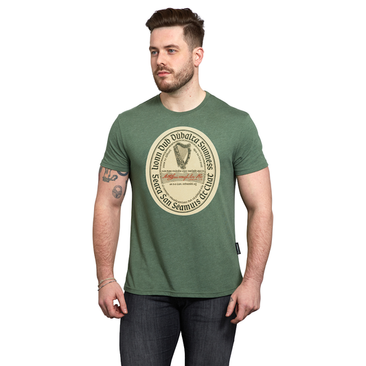 A man stands against a white background wearing the Guinness Green Distressed Label Tee, which features a vintage circular graphic and is made from a soft cotton blend for comfortable all-day wear.