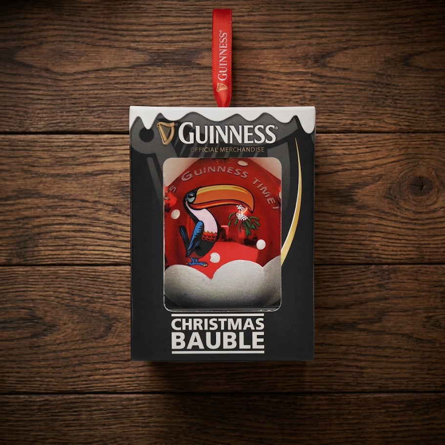 A boxed Guinness Toucan Glass Bauble with a striking red design featuring the iconic Guinness Toucan, snow, and the words “Guinness Time,” displayed on a wooden surface.