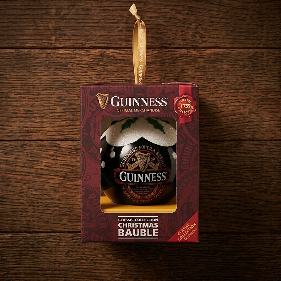 The Guinness Classic Label Glass Bauble comes in a festive box adorned with gold ribbon and iconic Guinness branding, displayed here on a wooden surface. This charming Christmas ornament is the perfect holiday gift for any beer lover.